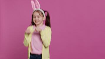 blij zorgeloos schoolmeisje jumping in de omgeving van in studio, imiteren een konijn en hoppen tegen roze achtergrond. vrolijk actief kind vervelend konijn oren en stuiteren, aanbiddelijk kind. camera a. video