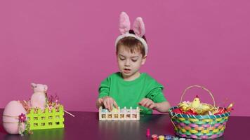 Happy pleased child decorating easter arrangements with handcrafted items, arranging painted colorful eggs in a basket. Joyful little boy celebrating spring festivity, pink backdrop. Camera A. video