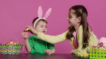 Sweet children knocking eggs together for easter tradition in studio, playing a seasonal holiday game against pink background. Lovely adorable kids having fun with festive decorations. Camera A. video