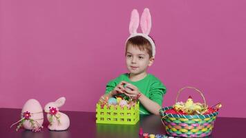 Excited little kid decorating baskets with eggs and grass to prepare for easter holiday festivity, creating festive arrangements. Cute toddler with bunny ears does craft activity. Camera A. video