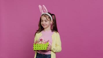 Smiling pleased girl presenting her handcrafted easter basket filled with painted eggs and other festive decorations for holiday celebration. Joyful small kid proud of her arrangement. Camera A. video