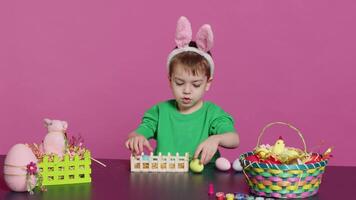 Happy toddler with bunny ears arranging basket filled with painted eggs, creating festive arrangements for the easter sunday celebration. Cheerful little kid enjoys art and craft. Camera A. video