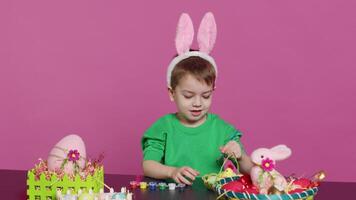 sorridente gioviale bambino in età prescolare pittura uova e ornamenti per Pasqua festa preparativi, utilizzando arte e mestiere materiale per decorare festivo disposizioni. giovane ragazzo avendo divertimento con cravatta tintura. telecamera un. video