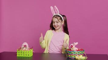 Happy schoolgirl with bunny ears showing thumbs up sign, feeling excited about easter holiday preparation. Cheerful child doing like okay sign and decorating baskets for festivity. Camera A. video