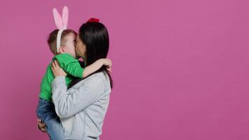 carino madre e bambino mostrando amore per ogni altro nel studio, mamma abbracciare e baci sua piccolo bambino piccolo figlio con coniglietto orecchie. adorabile poco famiglia ridendo insieme e avendo divertimento. telecamera un. video