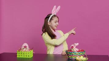 Adorable little kid indicating all directions in front of camera, pointing up, down, left and right while sitting at the table. Smiling lovely girl with bunny ears looking around. Camera A. video