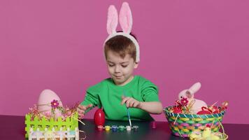 adorable pequeño joven crea hecho a mano adornos para Pascua de Resurrección domingo y pintura huevos con sellos y cepillos satisfecho joven chico utilizando Arte materiales a color y Decorar. cámara una. video