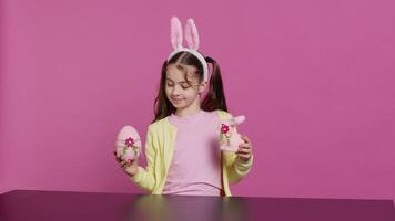 piccolo allegro ragazza mostrando colorato decorato ornamenti su telecamera, la creazione di fatto a mano festivo disposizioni. giovane bambino sensazione eccitato di Pasqua domenica, detiene coniglio e uovo. telecamera un. video