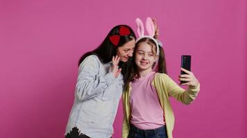 speels vrolijk moeder en meisje glimlachen voor foto's Aan smartphone, hebben pret omarmen elk andere in studio. weinig kind een haar mam poseren voor afbeeldingen, voor de gek houden in de omgeving van. camera b. video