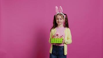 Smiling pleased girl presenting her handcrafted easter basket filled with painted eggs and other festive decorations for holiday celebration. Joyful small kid proud of her arrangement. Camera B. video