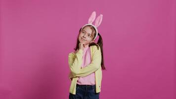glimlachen weinig meisje dromen over cadeaus en voedsel voor Pasen zondag viering, gevoel opgewonden over voorjaar vakantie. jong kind met konijn oren poseren tegen roze achtergrond. camera b. video