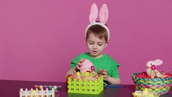 Joyful smiling boy decorating baskets and arrangements for easter sunday celebration, putting plastic grass and fake flowers to craft festive decorations. Happy toddler with bunny ears. Camera A. video