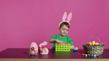 Sweet young boy making colorful arrangements for Easter holiday festivity, putting painted eggs in a handcrafted basket. Ecstatic joyful kid using crafting materials to create decorations. Camera B. video