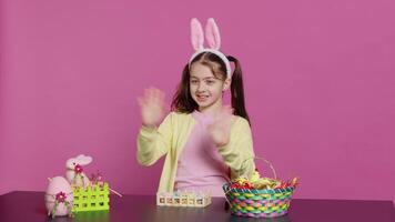 gioioso bambino mettendo coniglietto orecchie su sua testa e agitando a telecamera, detto Ciao e sorridente nel eccitazione per il Pasqua vacanza. contento bambino piccolo avendo divertimento con arte e mestiere attività. telecamera un. video