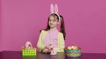 Cheery innocent child showing peace sign in studio and wearing bunny ears, celebrating easter sunday holiday with festive ornaments on a table. Happy energetic girl with creativity. Camera A. video