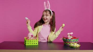 Enthusiastic young girl playing peek a boo in front of camera, using painted easter eggs against pink background. Joyful lovely toddler feeling excited about spring holiday festivity. Camera B. video