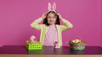 Smiling confident toddler placing bunny ears on her head and waving, saying hello against pink background. Cute joyful child creating decorations for easter sunday holiday event. Camera B. video