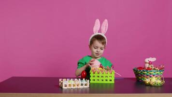 gioioso sorridente ragazzo decorazione cesti e accordi per Pasqua Domenica celebrazione, mettendo plastica erba e falso fiori per mestiere festivo decorazioni. contento bambino piccolo con coniglietto orecchie. telecamera b. video