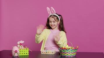 Joyful schoolgirl with bunny ears waving hello in front of camera, sitting at a table with festive decorations and arrangements for easter sunday celebration. Young excited child. Camera A. video