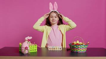 joyeux écolière avec lapin oreilles agitant Bonjour dans de face de caméra, séance à une table avec de fête décorations et arrangements pour Pâques dimanche fête. Jeune excité enfant. caméra b. video