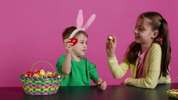 Sweet children knocking eggs together for easter tradition in studio, playing a seasonal holiday game against pink background. Lovely adorable kids having fun with festive decorations. Camera B. video