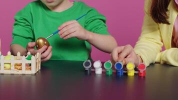 Playful children painting eggs using watercolor and brushes, coloring ornaments for easter sunday celebration. Brother and sister decorating items with stamps and art supplies. Camera A. video