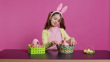 excité Jeune fille organiser peint des œufs dans une panier à préparer pour Pâques vacances fête, création de fête dispositions. espiègle content bambin avec lapin oreilles, Créatif activité. caméra b. video