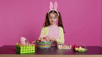 mignonne souriant bambin décorer une panier avec peint Pâques œufs, en présentant sa fabriqués à la main de fête arrangement dans studio. content charmant enfant création décorations et ornements. caméra b. video