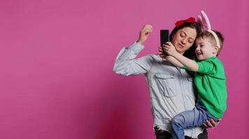 Lovely small child taking pictures with his mother on smartphone, trying to capture fun and cute moments against pink background. Little boy being playful and fooling around with phone. Camera B. video