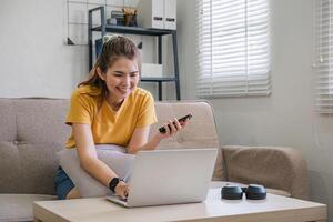 Happy young Asian woman uses mobile phone while sitting on sofa at home with laptop computer. photo