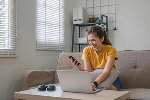 Happy young Asian woman uses mobile phone while sitting on sofa at home with laptop computer. photo