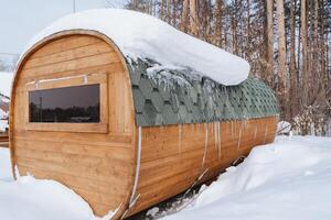 The bath barrel stands in nature under a layer of snow. Long wooden sauna made of boards. A window in a round house. Sauna in winter. photo