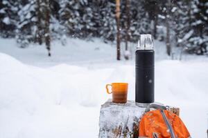 A black matte thermos, orange backpack for hiking and a mug with a spruce twig stand on a snow-covered stump.Warm tea in the winter frost.Minimalistic subject shot photo