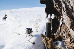 Thermos pan burner, camping camping utensils, old tree root, white snow, sunny weather. photo