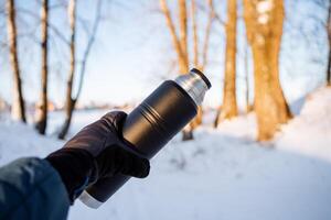 el mano sostiene un termo en contra el antecedentes de naturaleza. negro metal termo para té. mantener calentar en invierno. turista utensilios foto