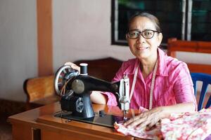 Happy senior Asian woman tailor is sewing cloth by vintage sewing machine at home. Concept, Senior lifestyle, spend free time to repair or design clothes. Hobby, not lonely. photo