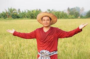 Handsome Asian man farmer wears hat, red shirt, raise arms open wide, stands at paddy field. feels happy. Concept, Agriculture occupation. Working with nature. Organic farming. photo