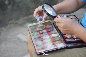 cerca arriba mano sostener aumentador vaso a inspeccionar detalle y modelo minúsculo de amuleto desde recopilación. concepto, Fé creencia en santo y suerte para budistas colectar amuleto como aficiones, intercambiar o comercio. foto