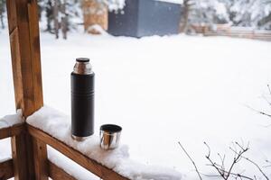 black matte thermos and it's metal mug with hot drink on window sill of the outdoor veranda. Hiking and picnic staff. Drinking warm tea or coffee in the winter frost. photo