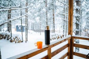 black matte thermos and orange mug with hot drink on window sill of the outdoor veranda. Hiking and picnic staff. Drinking warm tea or coffee in the winter frost. photo