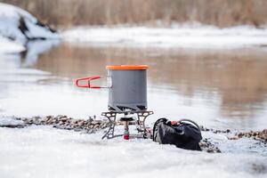 concept of tourist dishes, a pot with a lid stands on a gas burner on the bank of the river, boiling water in a hike in a pan. winter hike in the mountains. photo