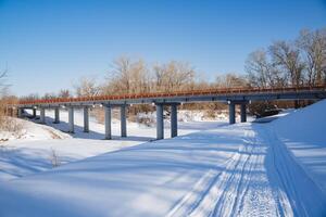 A bridge over a frozen river against a background of white snow. A road across a bridge in a remote forest. Snowmobile footprints in the snow. Winter landscape. photo