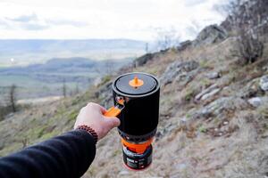 A man's hand holds a pot for eating against the backdrop of mountains, a fast food system, a safe gas burner, hiking equipment, trekking utensils. photo