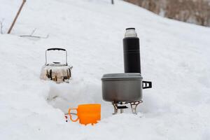 Cooking utensils for hiking, tea mug, hot drink thermos, pot on burner, kettle in the snow, winter tourism cold in the forest, breakfast in nature without people photo