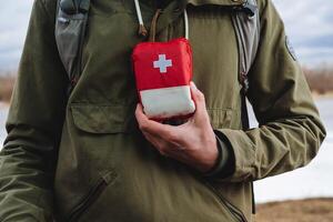 un turista persona sostiene en su mano un primero ayuda equipo, un rojo médico bolso para emergencias, salud cuidado, un blanco cruz, cámping equipo. foto