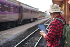 Asian man traveller is at railway station, holds smart tablet. Concept, travel by train in Thailand can book or buy ticket, check timetable online. Technology transportation. photo