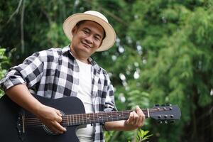 Handsome Asian man is playing acoustic guitar in the park, outdoor nature background. Concept, love music, hobby, recreation activity. Relax time. Music can develop mental health. photo