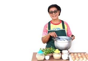 Happy Asian senior woman is cooking, wear chef cap and apron, holds pot and ladle. Concept, Cooking for family. Cooking for sell. Thai kitchen lifestyle. Elderly activity. photo