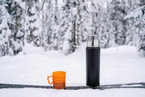 black matte thermos and orange mug with coffee and tea in a snow. Hiking and picnic staff. Drinking warm tea in the winter frost. Minimalistic subject shot photo