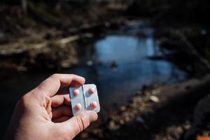 The hand holds the tablets against the background of the river. A man took medicine in the forest. Pink pills. Emergency assistance in the hike. photo